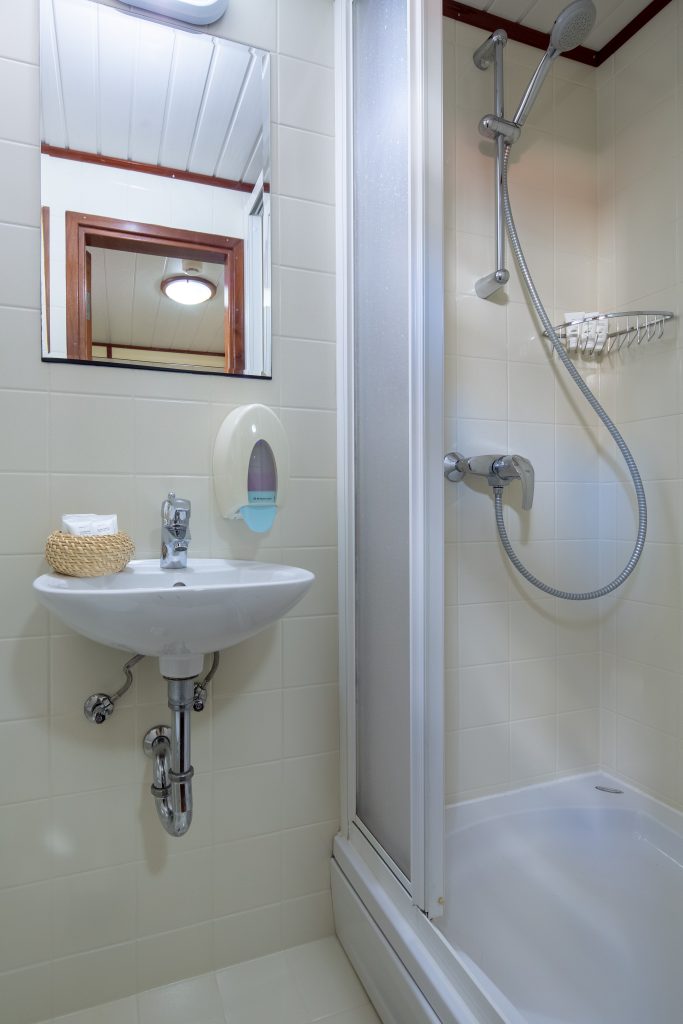 A small bathroom on a yacht charter with a wall-mounted sink, soap dispenser, mirror, and a corner shower featuring a sliding door, showerhead, and wire shelf. The white walls and fixtures offer a clean, modern look for your yacht rental experience.