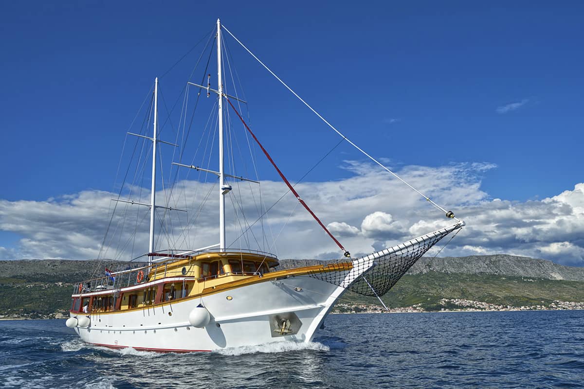 A large white sailboat with wooden trim sails on a calm blue sea under a clear sky, with mountains and a coastal town visible in the background.
