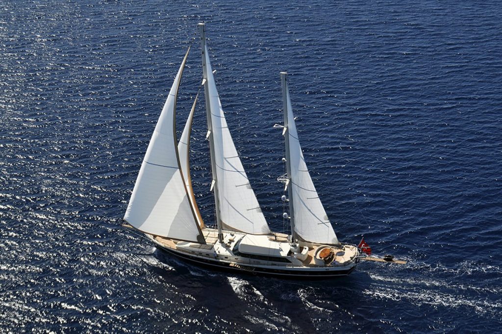 A large sailboat with three white sails glides on deep blue water, leaving a small wake behind it. The boat has a wooden deck and a red flag at the stern. The sun glints off the waves.