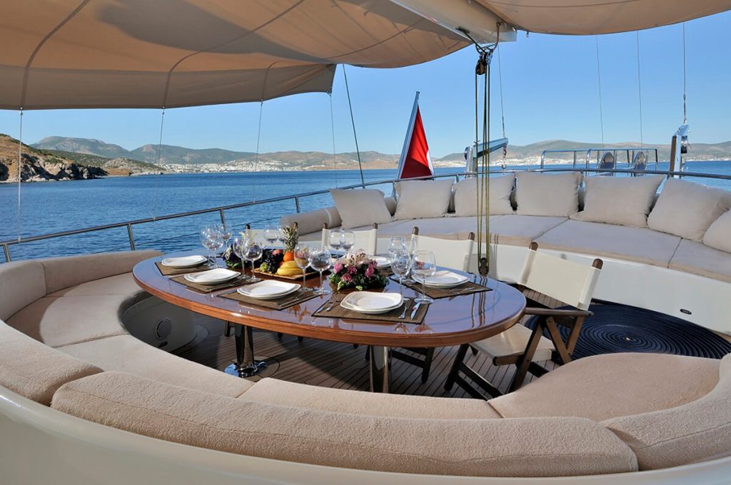 A luxury yacht deck with a curved sofa, an elegant dining table set for six, and scenic views of the sea and distant coastline under a clear blue sky.