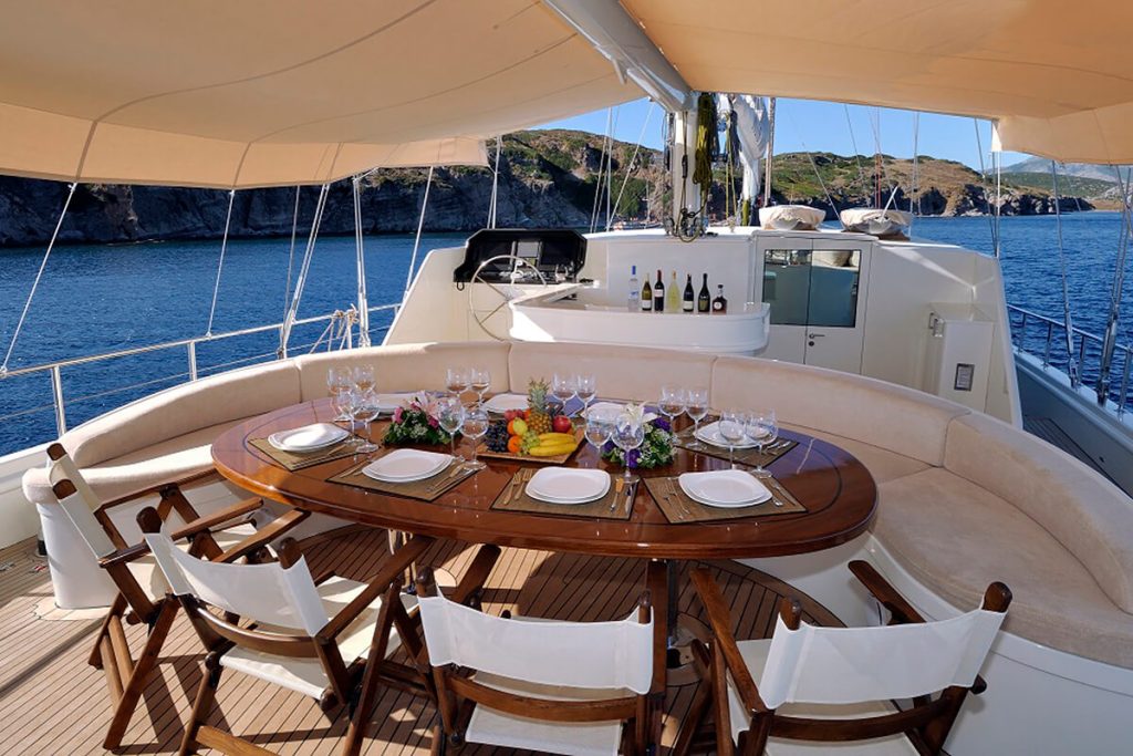 A yacht deck with a shaded dining area featuring a wooden table set for six, decorated with fruit and flowers. Sea and cliffs are visible in the background, with a bar and bottles near the yacht’s helm.