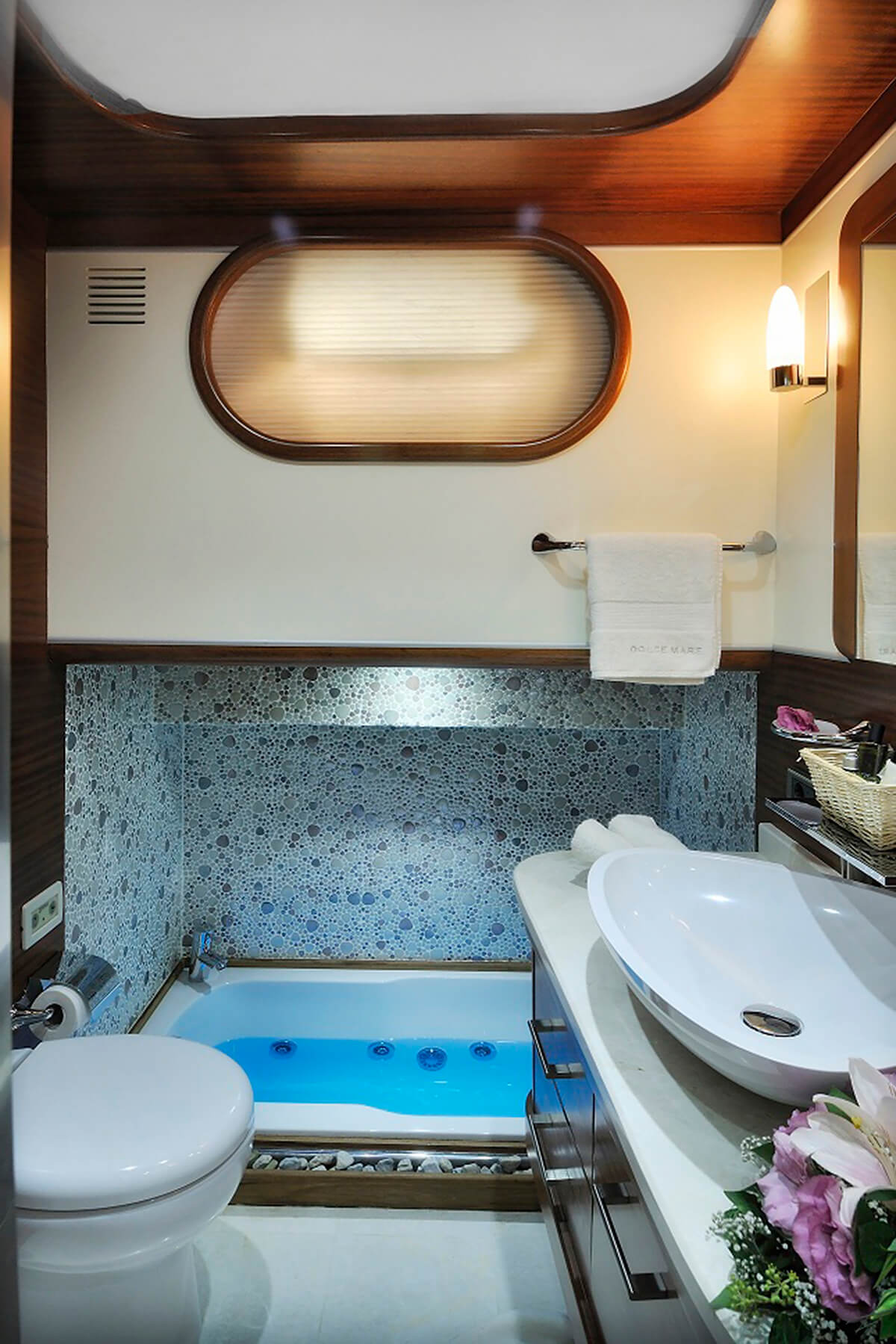 A modern bathroom with a circular window, mosaic-tiled sunken bathtub with blue water, wall-mounted towel rack, white toilet, vessel sink on a counter, and decorative flowers beside the sink.