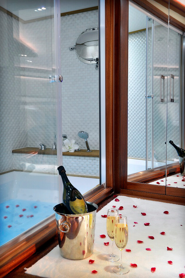 A luxurious bathroom with a bathtub, glass doors, and rose petals on the counter. A champagne bottle in an ice bucket and two filled flutes sit on a white towel beside the tub.
