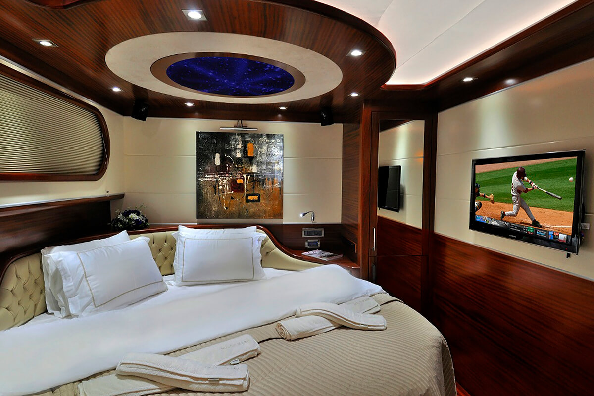 Luxurious bedroom on a yacht with wood paneling, a large bed with white linens, a TV showing a baseball game, modern artwork on the wall, and a starry ceiling light feature above the bed.