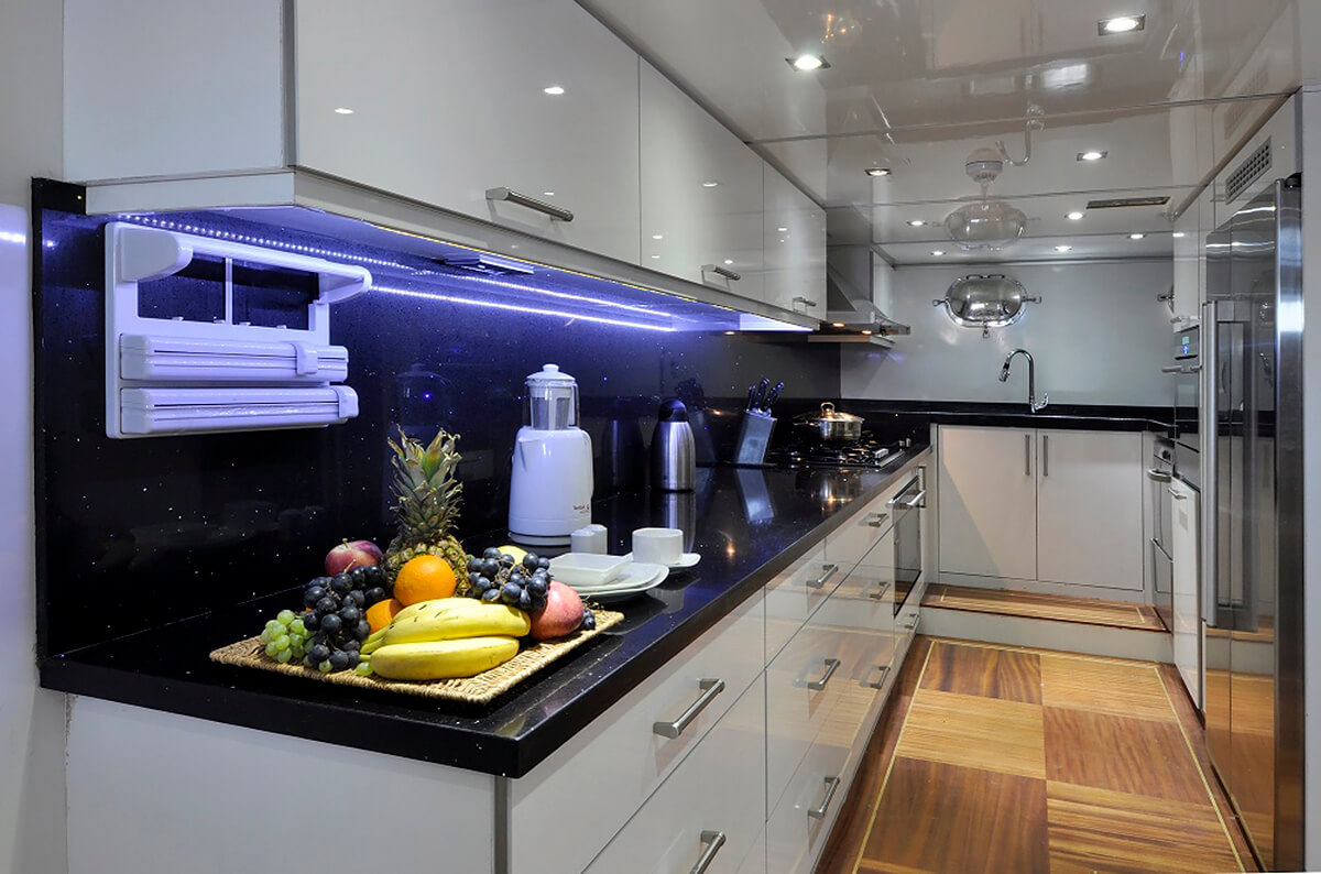 Modern kitchen with glossy white cabinets, black countertops, and stainless steel appliances. A tray of assorted fruits sits on the counter beside a blender, kettle, and under-cabinet paper towel holder.