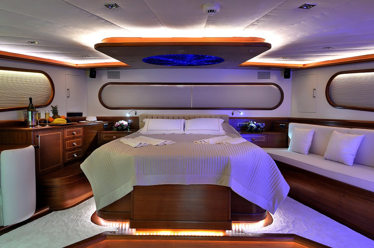 Luxurious yacht bedroom with a large bed, elegant wood paneling, ambient lighting, curved sofa seating, and a table with wine, fruit, and flowers, creating a cozy and upscale atmosphere.
