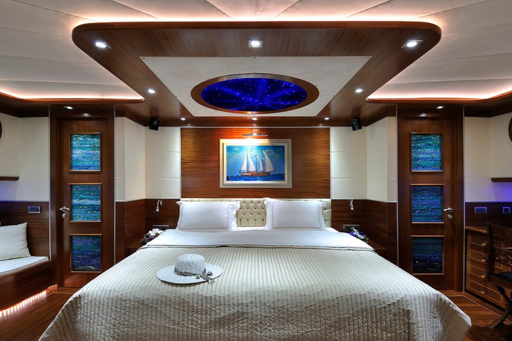 Luxurious bedroom with a large bed, white and beige bedding, a hat on the duvet, wood-paneled walls, marine-themed art, vibrant blue ceiling lights, and soft ambient lighting.