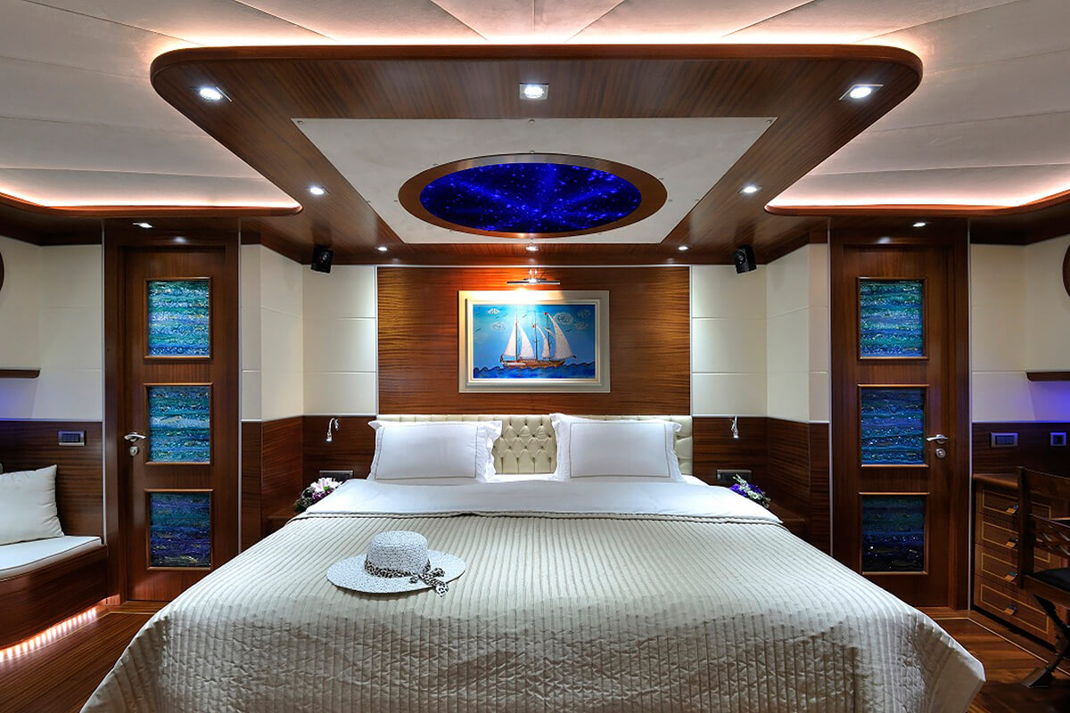 Luxurious bedroom with a large bed, white and beige bedding, a hat on the duvet, wood-paneled walls, marine-themed art, vibrant blue ceiling lights, and soft ambient lighting.