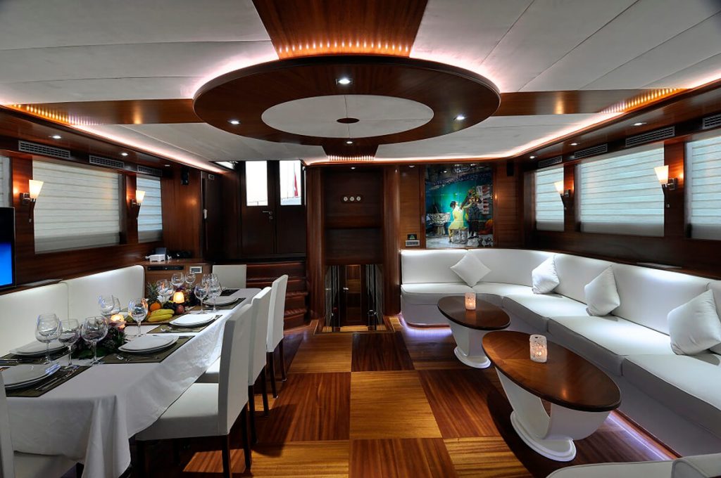 Luxurious yacht interior featuring a dining table set for dinner, a curved white sofa with cushions, wooden flooring and ceilings, soft ambient lighting, and decorative candles on small round tables.