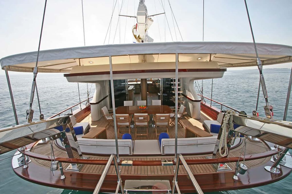 The image shows the deck of a luxury yacht with cushioned seating, a dining table with chairs, and open space overlooking calm blue sea under a covered canopy.