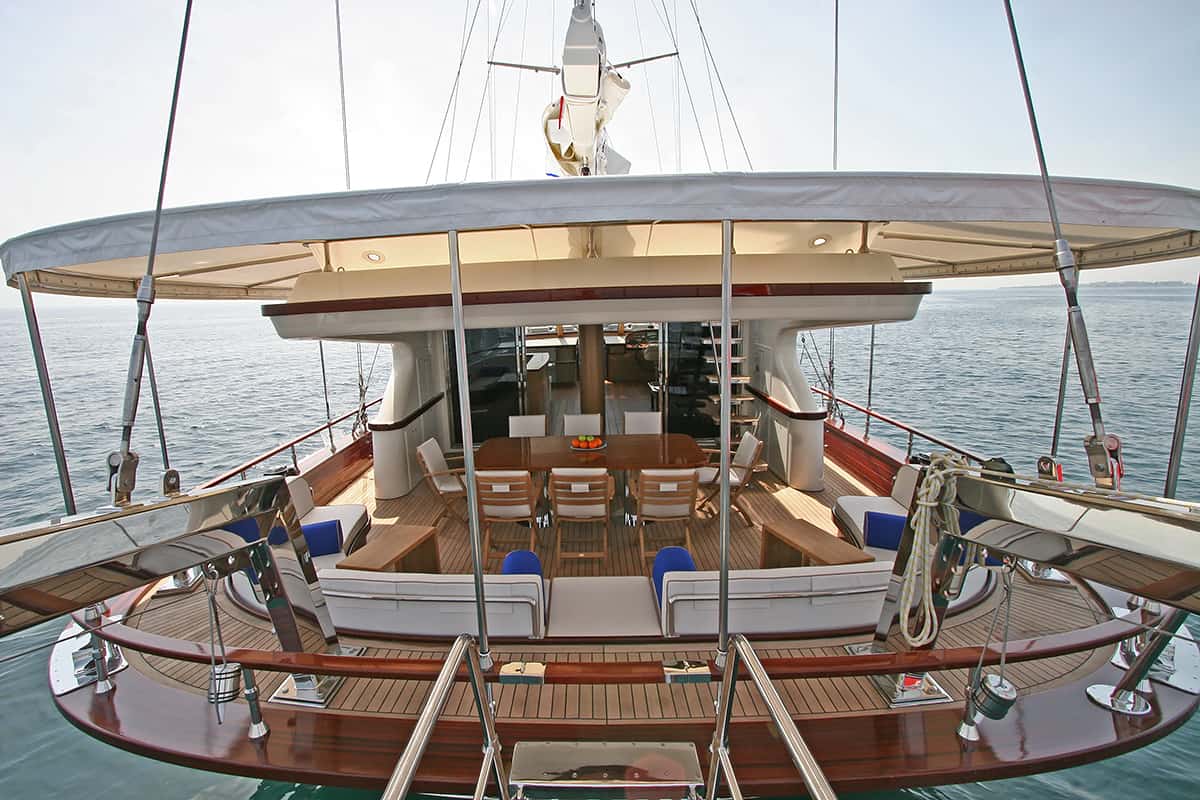The image shows the deck of a luxury yacht with cushioned seating, a dining table with chairs, and open space overlooking calm blue sea under a covered canopy.