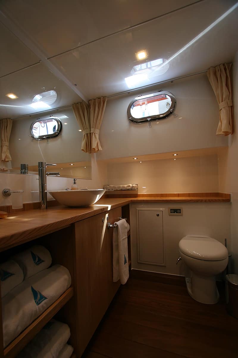 Modern boat bathroom with round porthole windows, wooden countertop, vessel sink, mirror, toilet, shelves with folded towels underneath, and beige curtains. Warm lighting creates a cozy atmosphere.