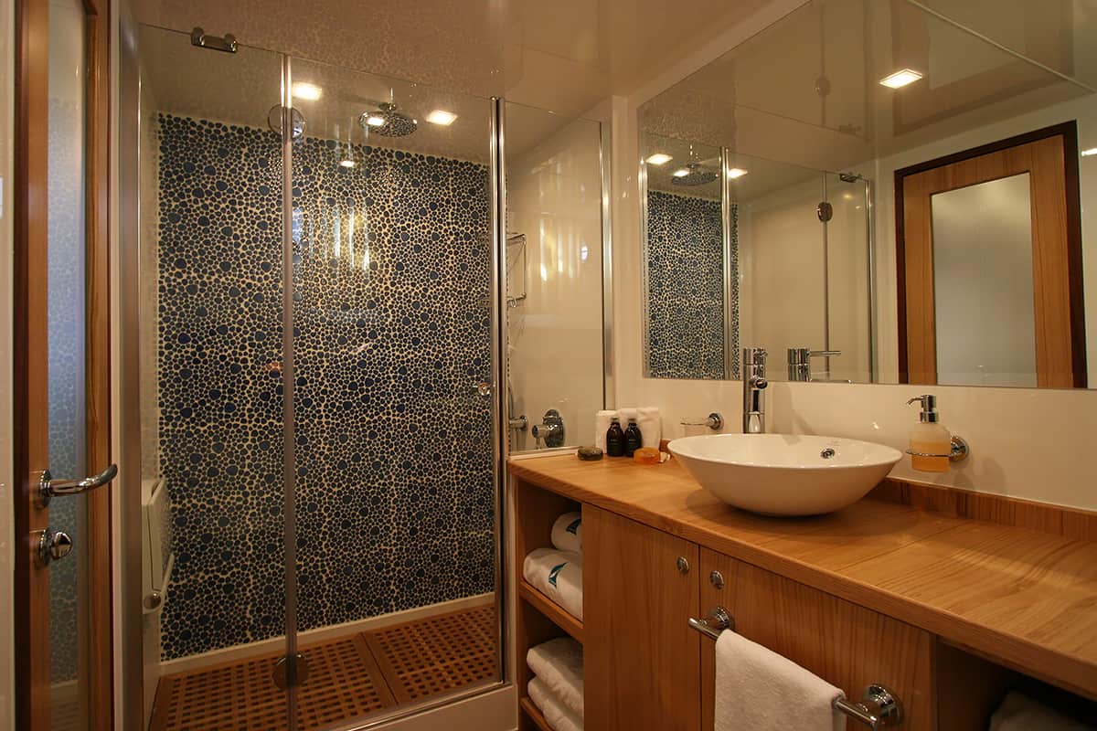 Modern bathroom with a glass-enclosed shower featuring a pebble tile wall, a vessel sink on a wooden countertop with storage shelves, mirror, and various toiletries neatly arranged.
