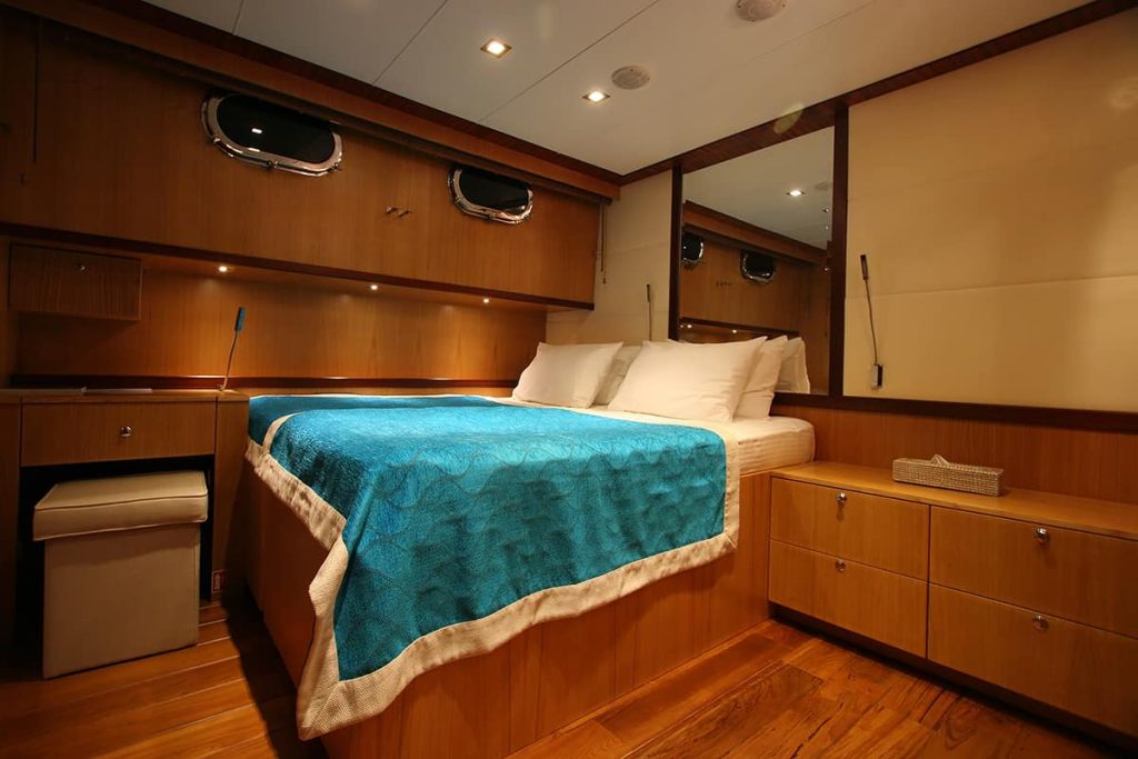 A cozy bedroom with wood-paneled walls and furniture, a bed with white pillows and a blue blanket, two small porthole windows, built-in drawers, a mirror, and warm overhead lighting.