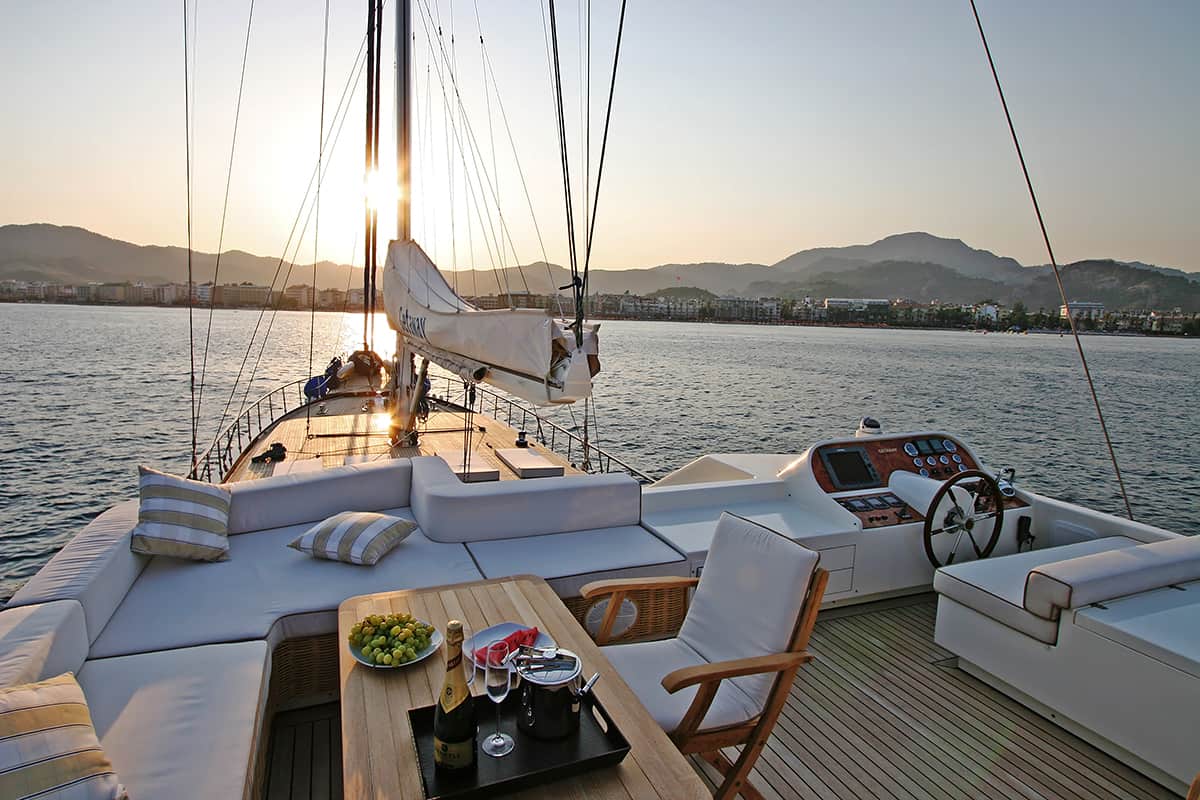 A luxury yacht deck at sunset features cushioned seating, a wooden table with wine and snacks, and a steering wheel, with calm water and mountains in the background.