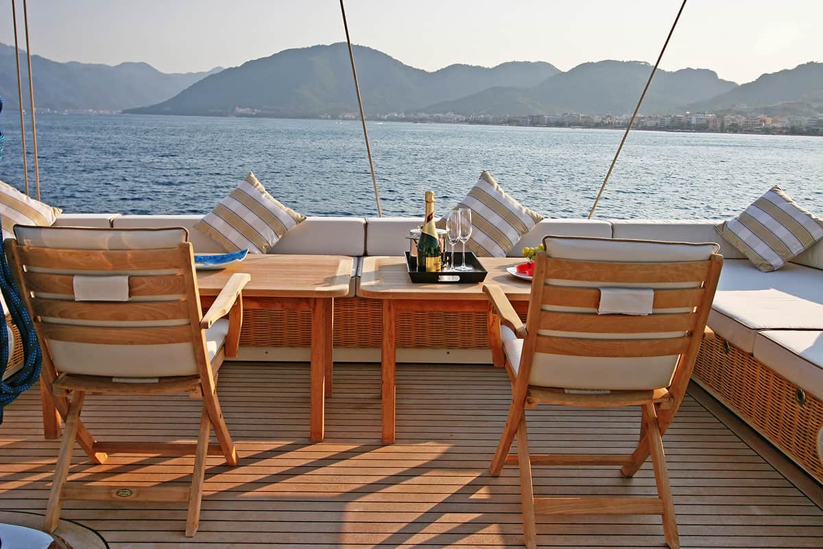 Two wooden chairs and a table with wine and glasses are set on a yacht deck with cushioned seating. The yacht overlooks calm blue water and distant green mountains under a clear sky.