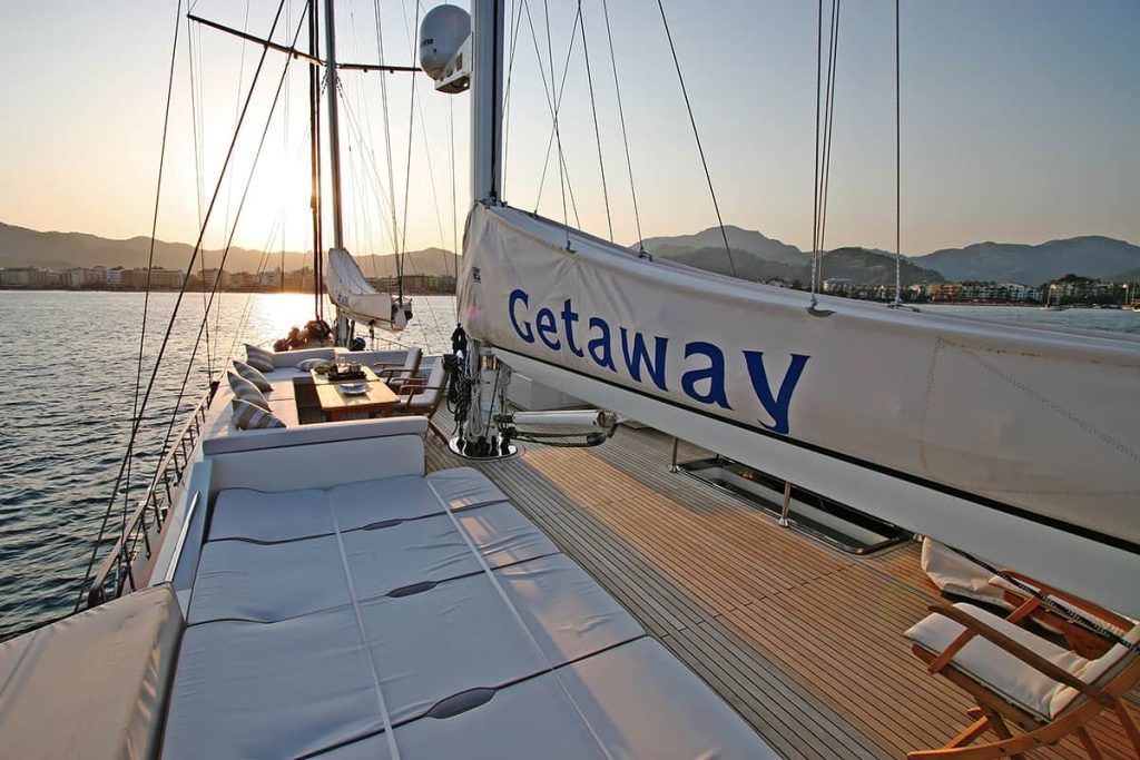 A luxury sailboat named Getaway on calm water at sunset, with mountains and a coastal city in the background and lounge seating on the spacious deck.