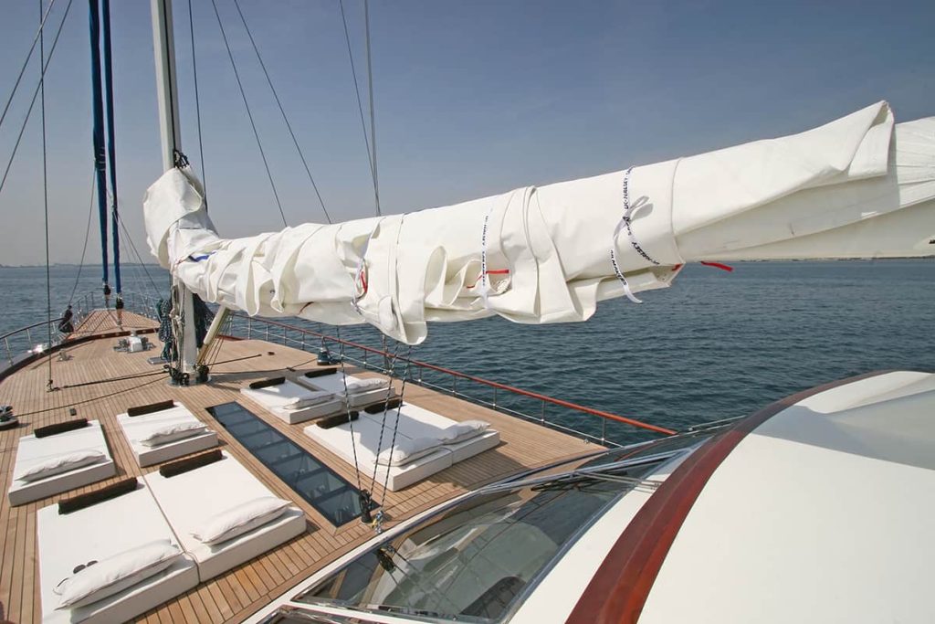 View from the deck of a modern sailboat with a wrapped sail, lounge cushions, and clear blue water in the background under a sunny sky.