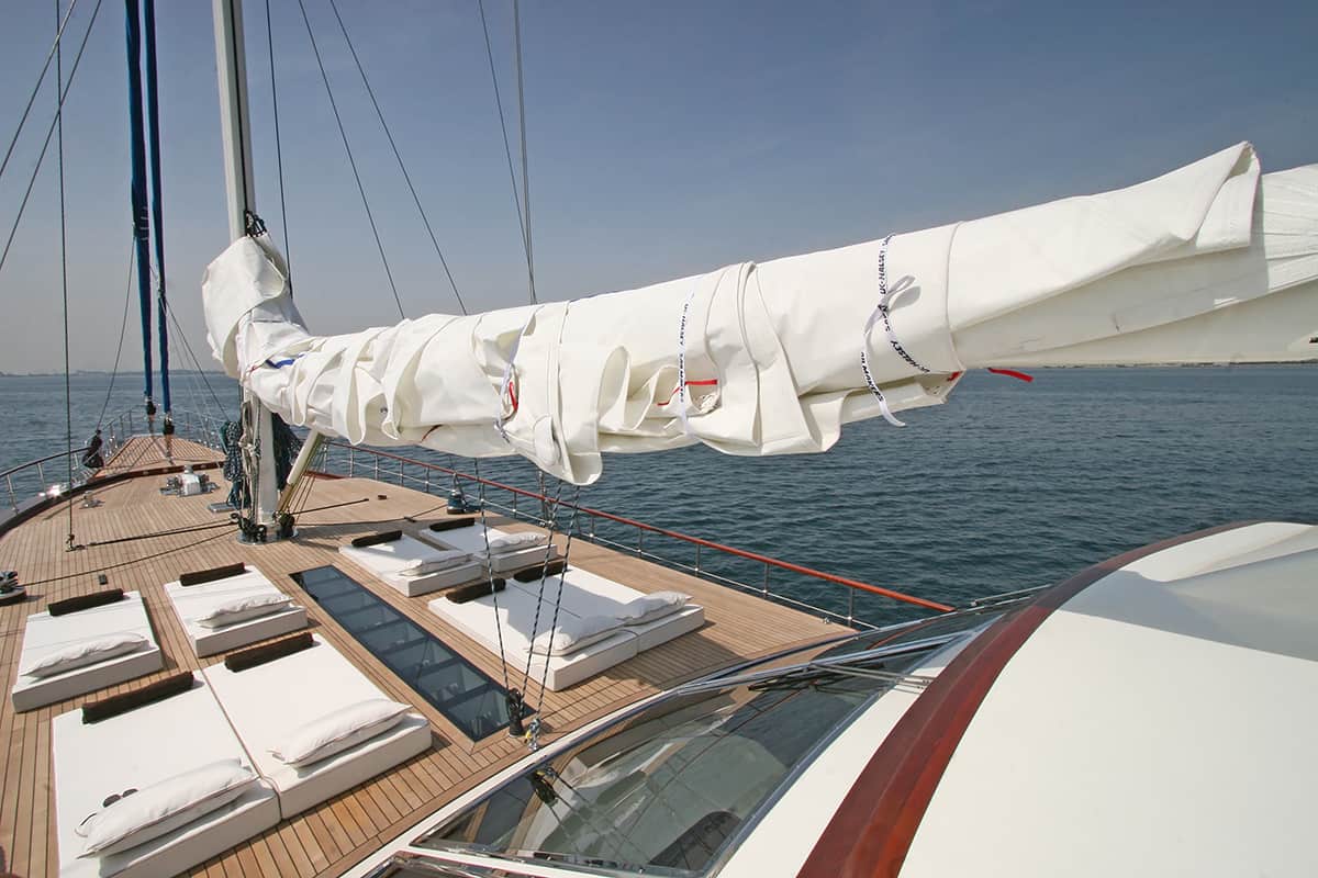 View from the deck of a modern sailboat with a wrapped sail, lounge cushions, and clear blue water in the background under a sunny sky.