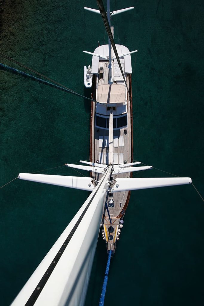 Aerial view looking straight down from the mast of a sailboat anchored in clear, turquoise water, showing the deck, masts, and a small dinghy tied alongside.
