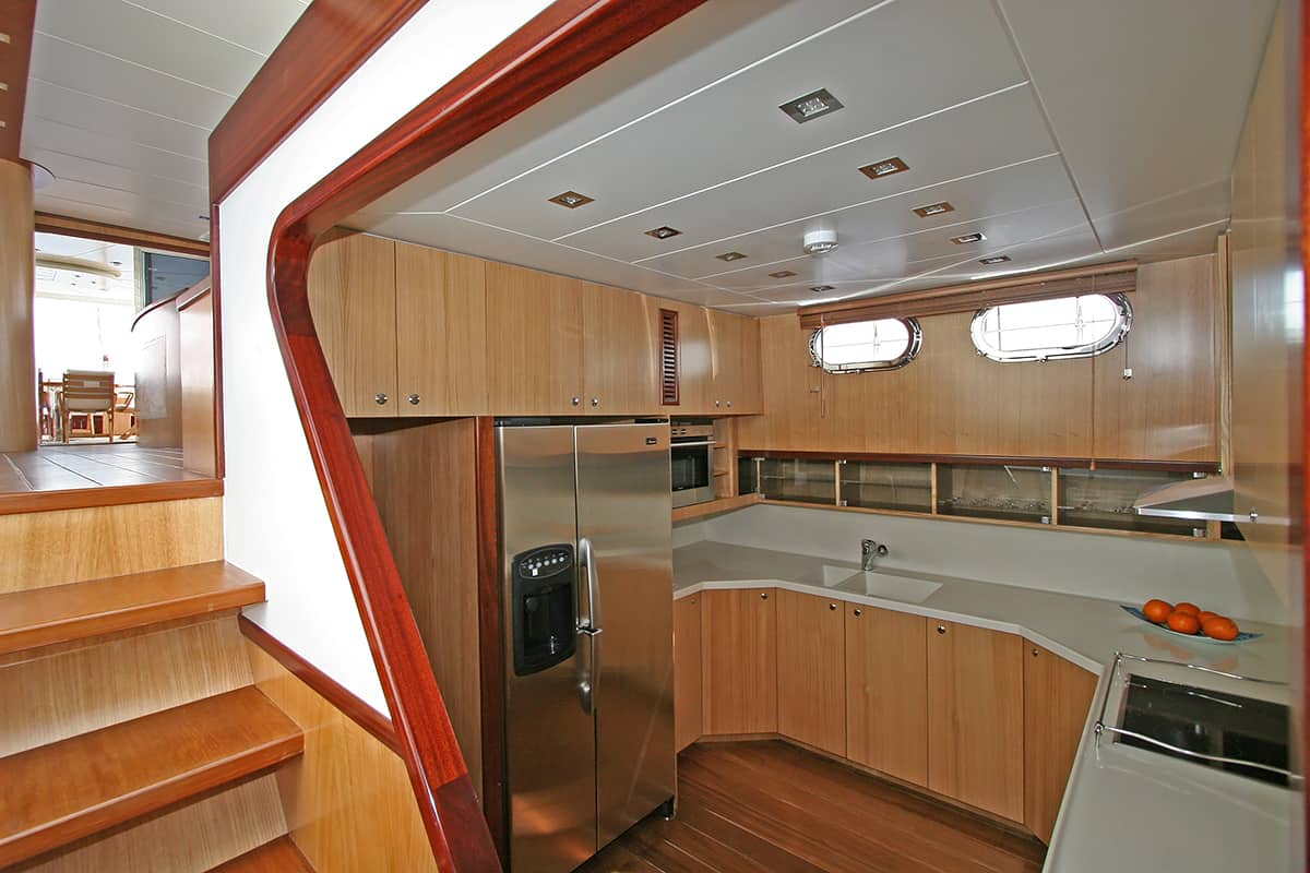 Modern yacht kitchen with wooden cabinets, stainless steel refrigerator, sink, stove, and porthole windows. A bowl of oranges sits on the counter, and wooden stairs lead up to another area.