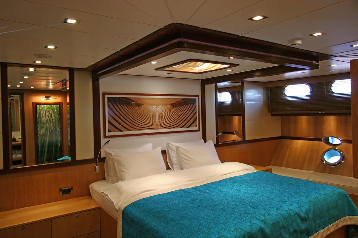 Luxurious yacht cabin with a double bed, teal blanket, white pillows, wooden paneling, framed artwork above the bed, mirrors, and round porthole windows letting in natural light.