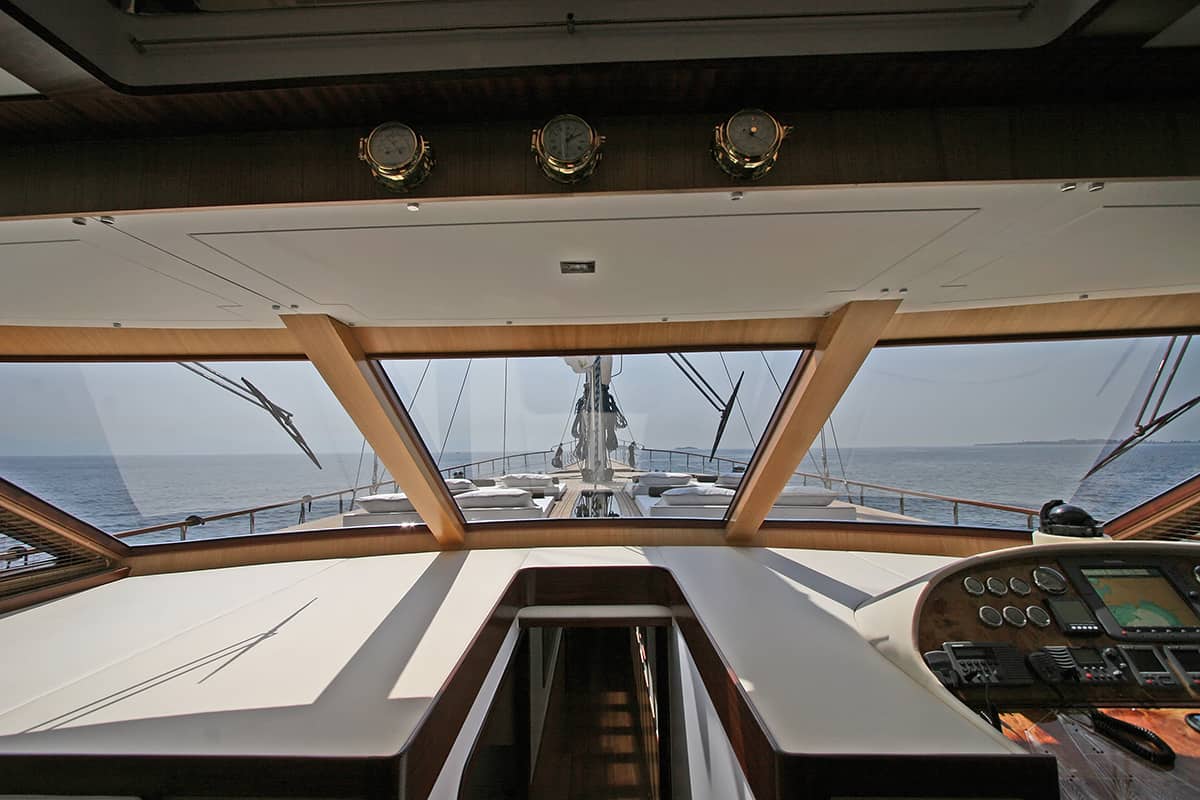 View from inside a yacht’s cockpit showing navigation instruments, a control panel, and large windows overlooking the deck and calm ocean under a clear sky.