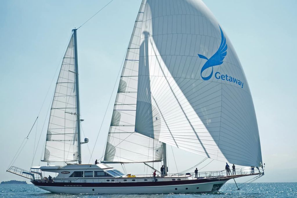 A large white sailboat named “Getaway” sails on calm blue water under a clear sky, with several people on board. The main sail features a blue bird logo and the word Getaway.