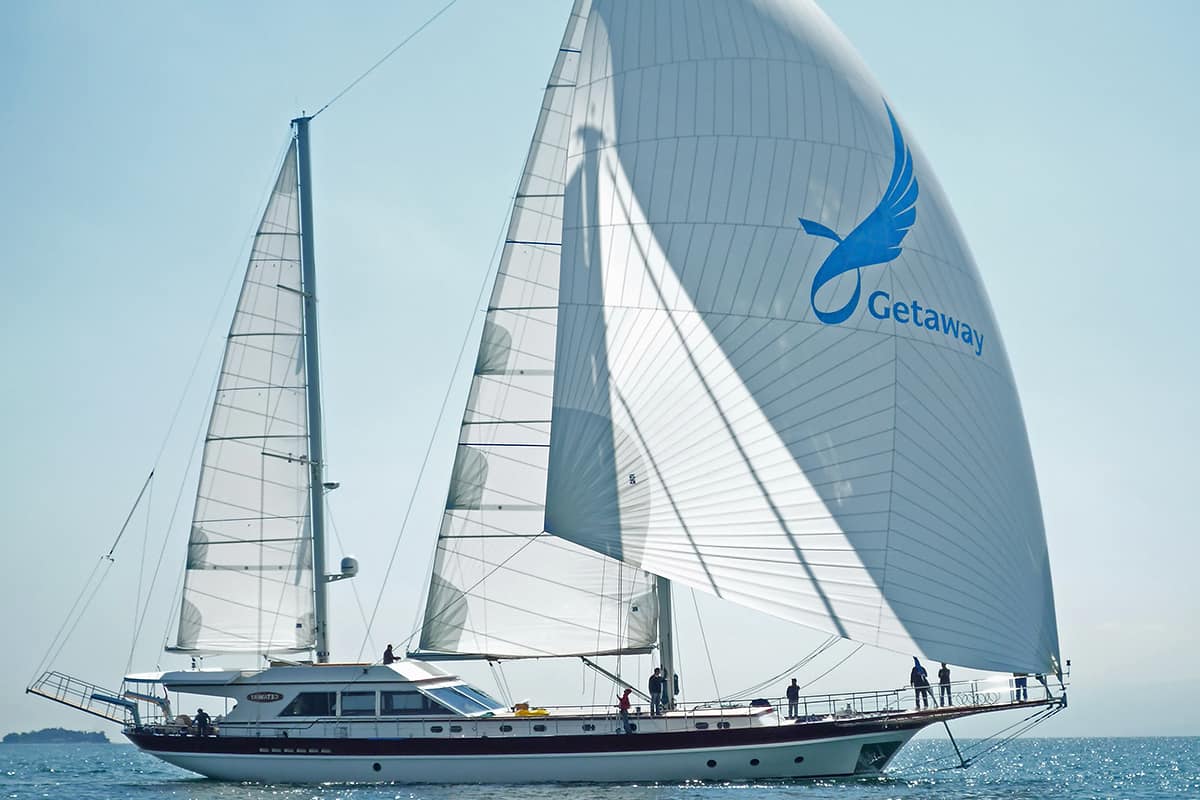 A large white sailboat named “Getaway” sails on calm blue water under a clear sky, with several people on board. The main sail features a blue bird logo and the word Getaway.