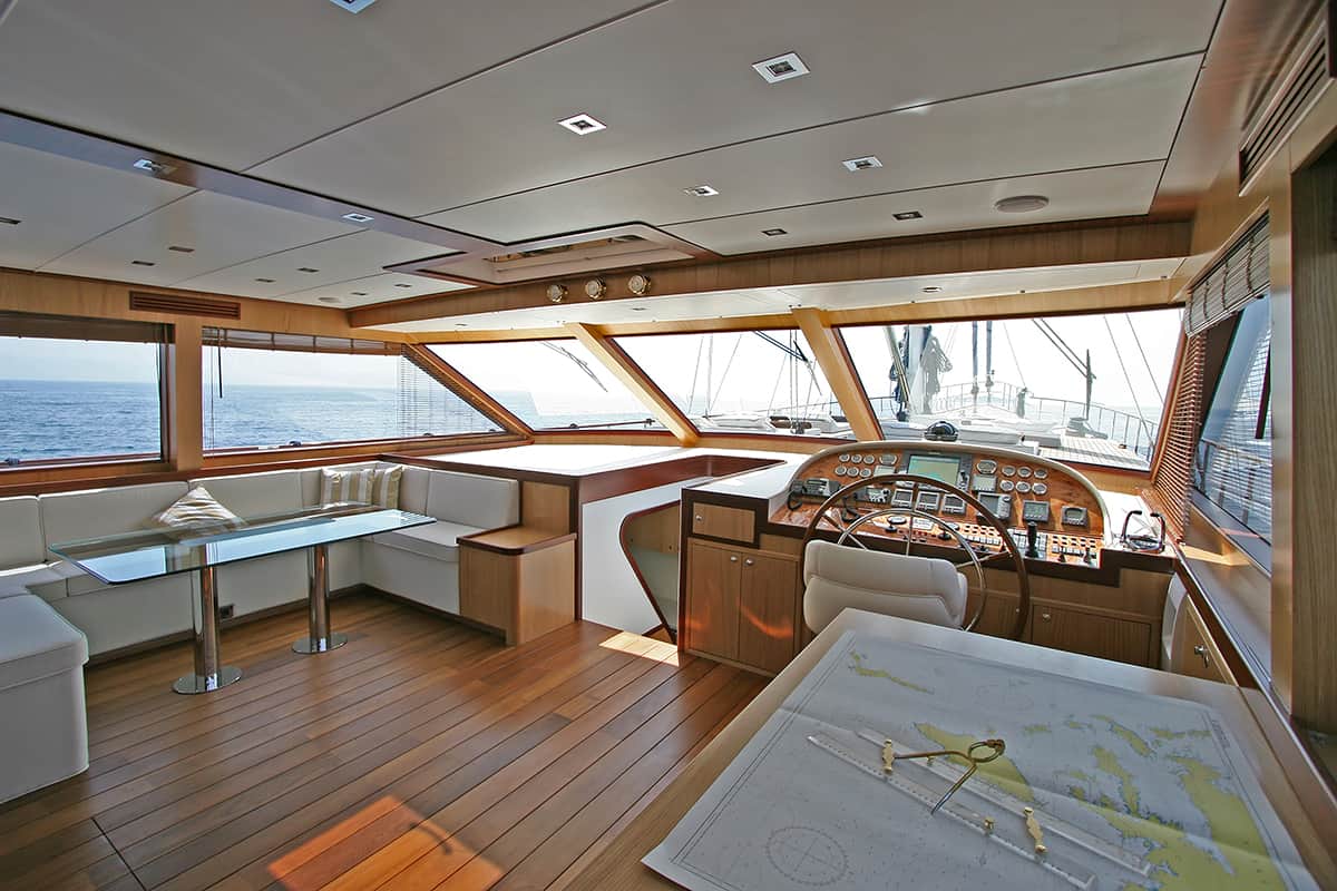 The interior of a yacht’s helm with large windows overlooking the sea, a captain’s control panel, a wooden floor, a dining area with white seating, and a navigation chart on a table in the foreground.