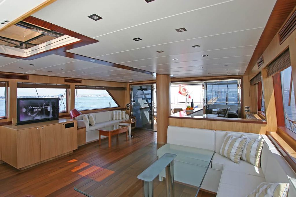 Modern yacht interior with wood floors and white furniture, large windows showing water views, a flat-screen TV, glass coffee table, and striped pillows; natural light fills the spacious, elegant area.