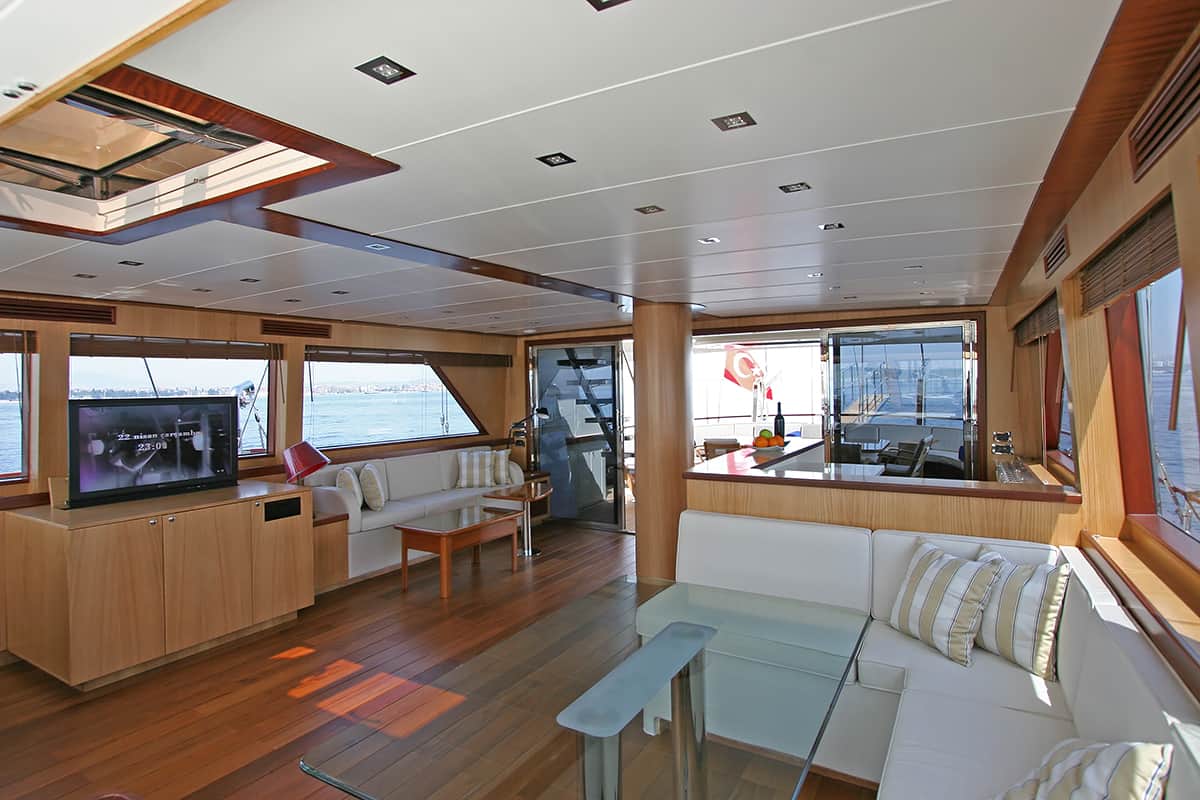 Modern yacht interior with wood floors and white furniture, large windows showing water views, a flat-screen TV, glass coffee table, and striped pillows; natural light fills the spacious, elegant area.