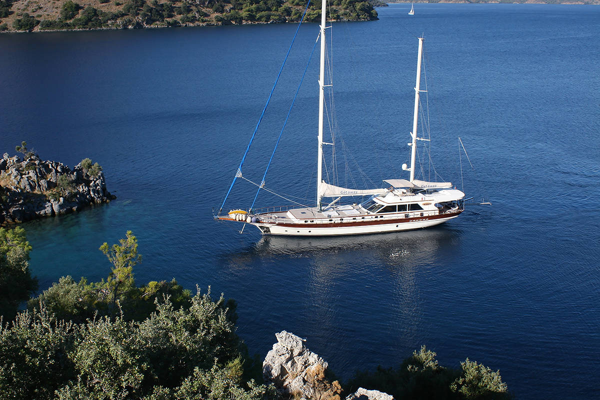 A white and brown sailboat with two masts floats on calm blue water near a rocky shore with green trees and shrubs, under clear skies.