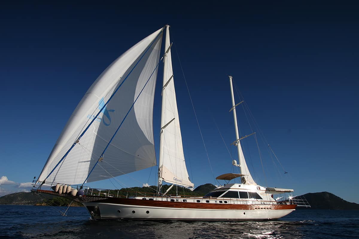 A large white sailing yacht with tall masts and fully unfurled sails glides on calm blue water, with green hills and a clear, deep blue sky in the background.