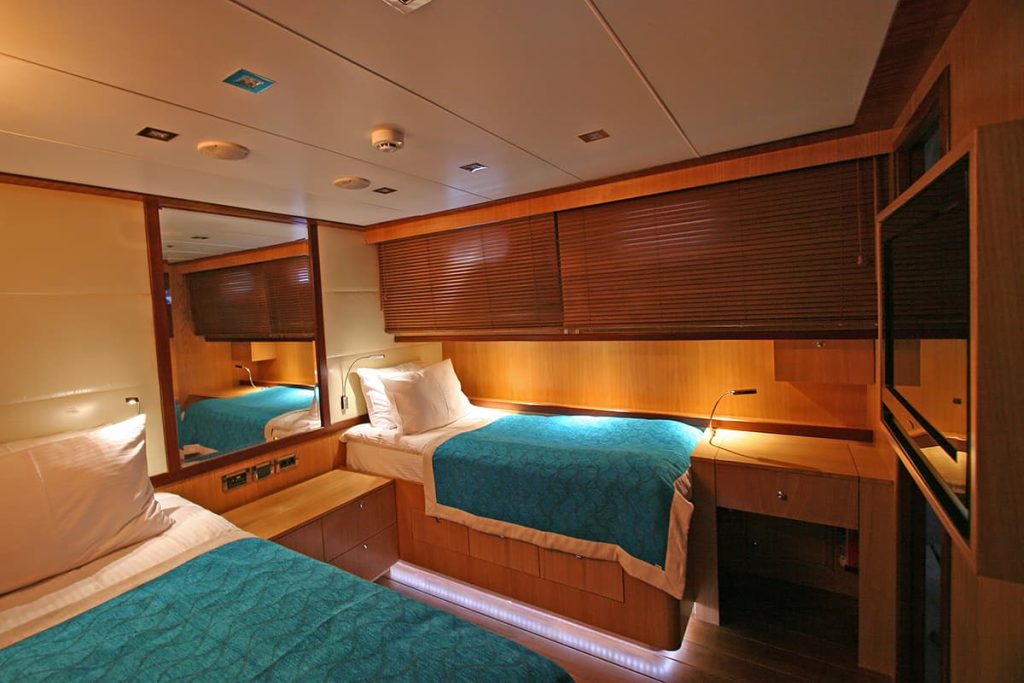 A cozy yacht cabin features two single beds with teal bedspreads, wood-paneled walls, built-in shelves, a large mirror, and a window with closed blinds. Warm lighting creates a comfortable atmosphere.