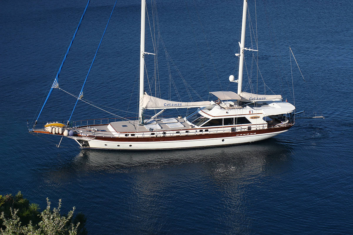 A large white sailing yacht with two masts, named Getaway, is anchored on calm blue water near the shoreline, with some greenery visible in the corner of the image.