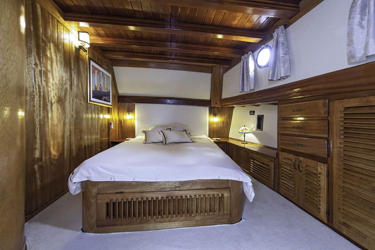 A cozy bedroom with wooden walls and ceiling, a double bed with white bedding, wall-mounted lights, small curtains on porthole windows, storage cabinets, and a framed picture on the wall.