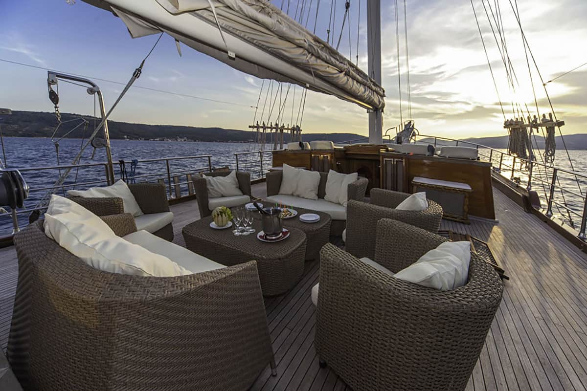 Wicker chairs with cushions and a table set with food and drinks are arranged on the deck of a sailboat at sunset, overlooking calm water and distant hills.