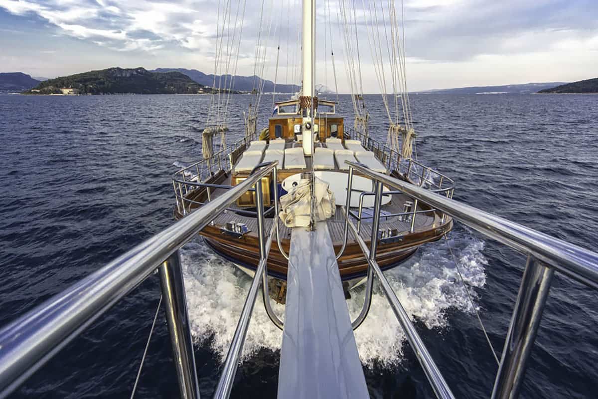 A view from the front of a wooden sailboat cruising on blue water, with waves splashing at the bow and hills visible in the background under a partly cloudy sky.