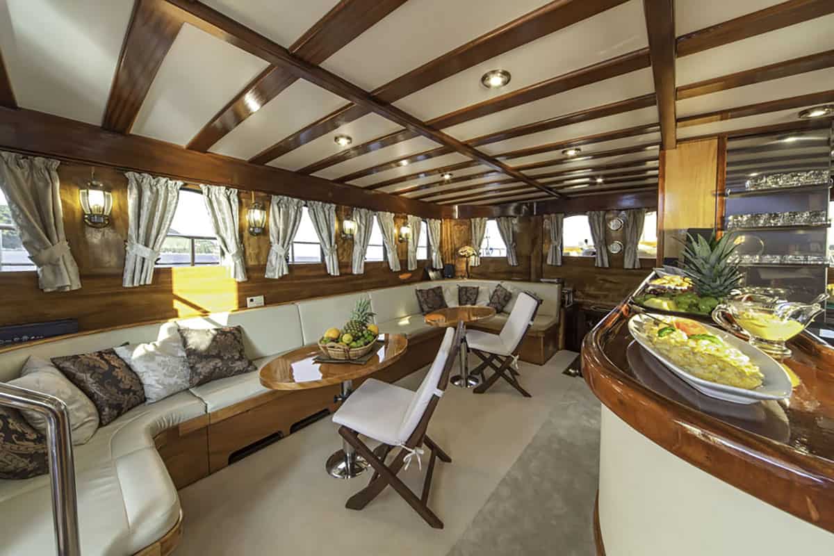 Luxurious yacht interior with wood-beamed ceiling, curved cream sofa, dining table, cushioned chairs, and a bar with plates of food. Sunlight streams through curtained windows, creating a warm and inviting atmosphere.