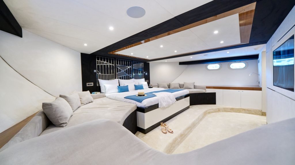 Luxurious yacht bedroom with a large bed, blue and white bedding, beige sofa, soft lighting, small windows, and sandals on the floor. The space is modern with light-colored walls and wooden accents.