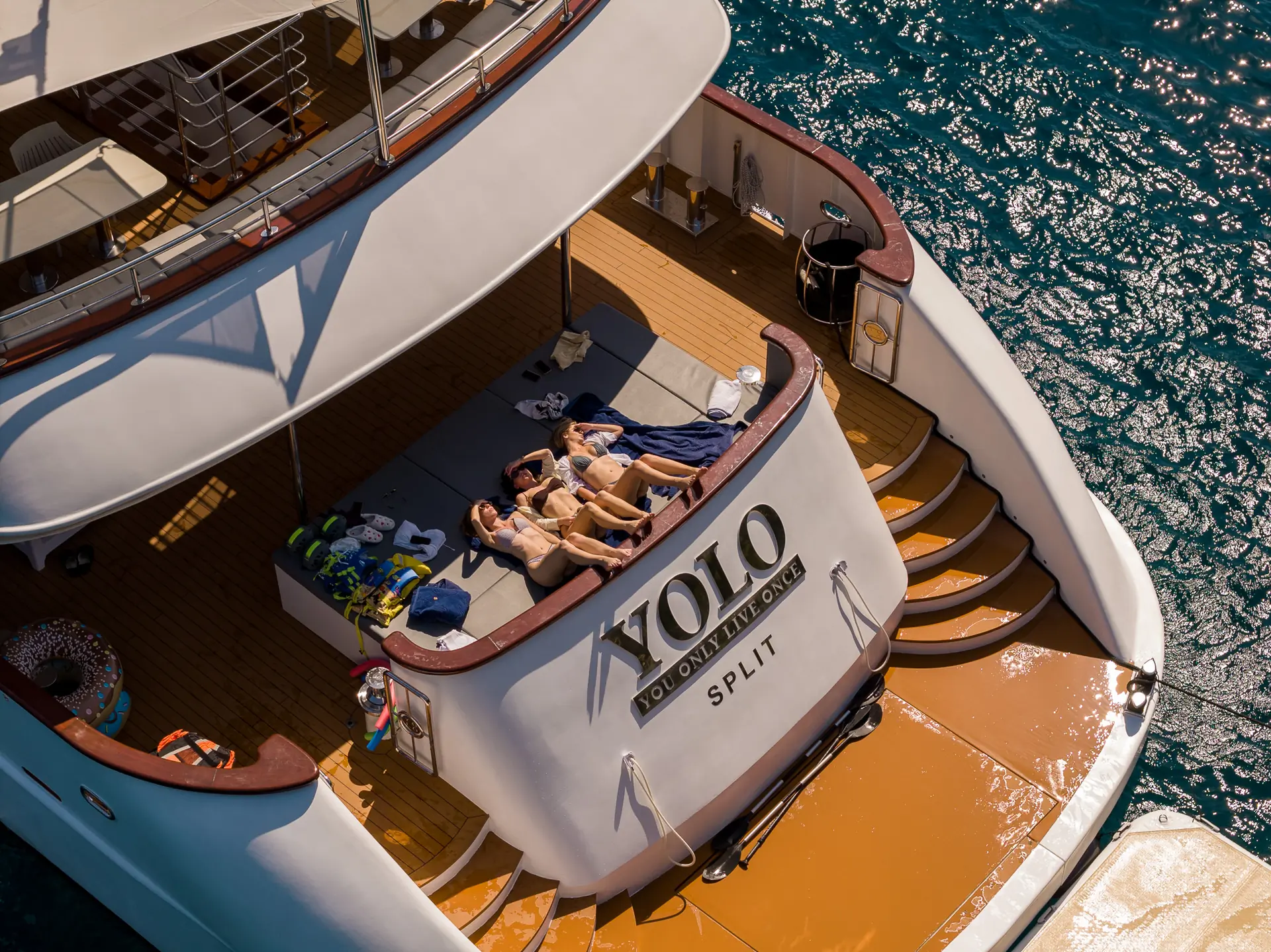 Aerial view of three people sunbathing on the back deck of a yacht named YOLO, docked in clear blue water. Towels, clothes, and water gear are scattered nearby on the wooden deck.