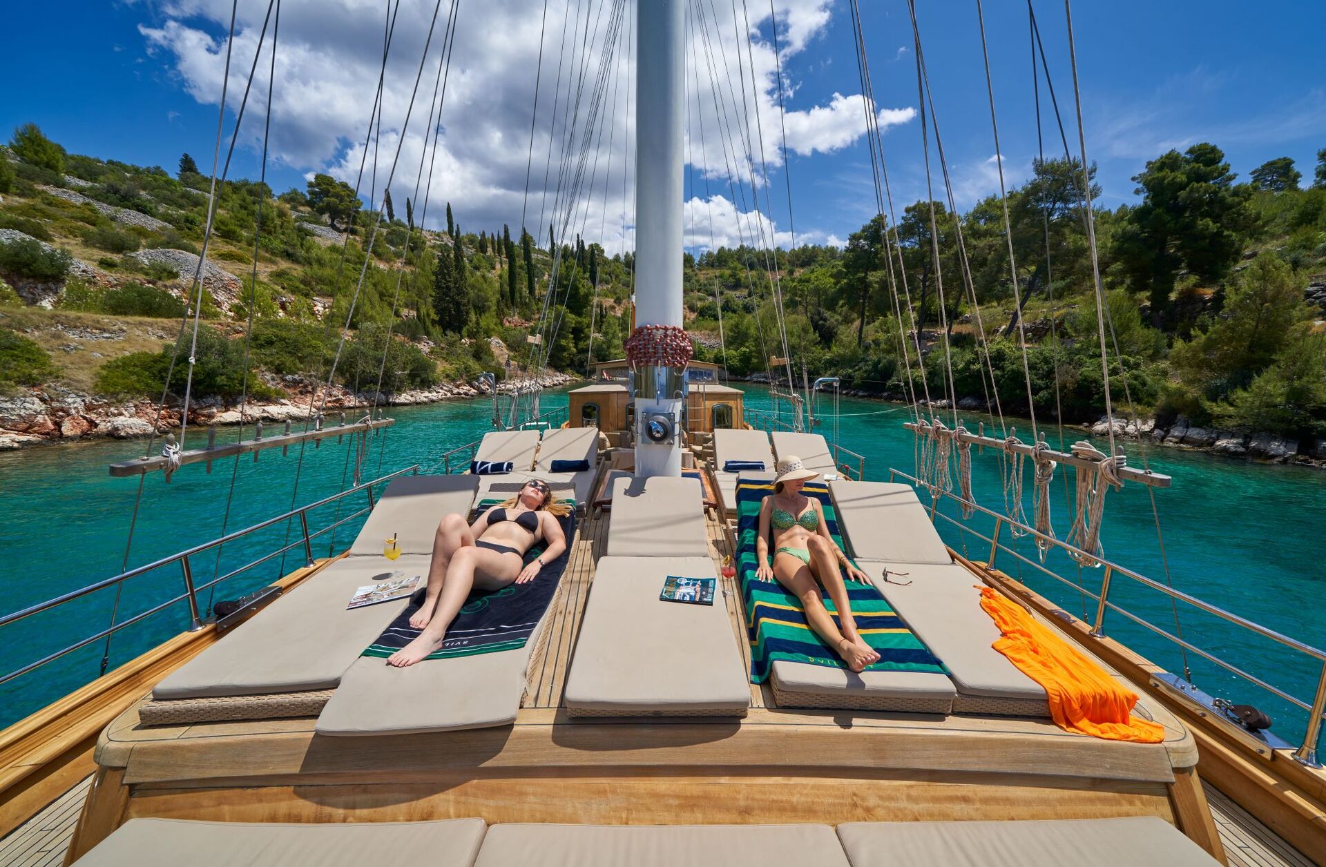 LIBRA Two women in swimsuits relax on sun loungers atop a yacht deck, surrounded by clear turquoise water and lush green trees under a sunny, partly cloudy sky.