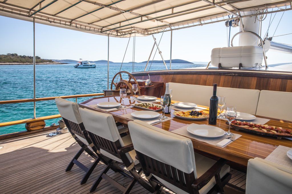 A set dining table with wine and food is on the deck of a yacht, overlooking calm blue water and distant hills under a sunny sky. Another yacht is visible in the background.