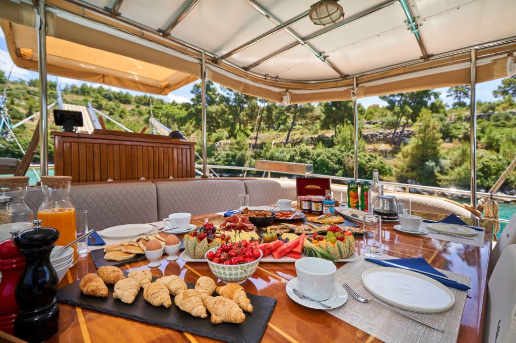 LIBRA A breakfast spread with croissants, fruit, eggs, and juice is set on a wooden table aboard a boat, with trees and greenery visible outside under a sunlit canopy.