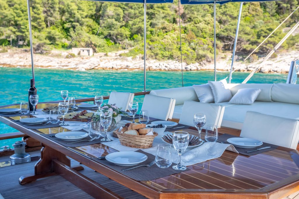 A dining table set with plates, glasses, wine, bread, and snacks on a yacht deck, with cushioned seating and a blue canopy, overlooking turquoise water and a forested shoreline.