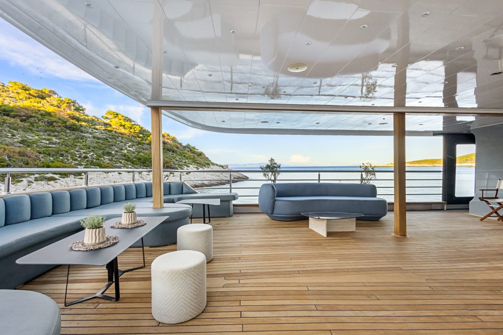 A spacious yacht deck with modern gray seating, small tables, potted plants, and wood flooring overlooks calm water and tree-covered hills on a sunny day.