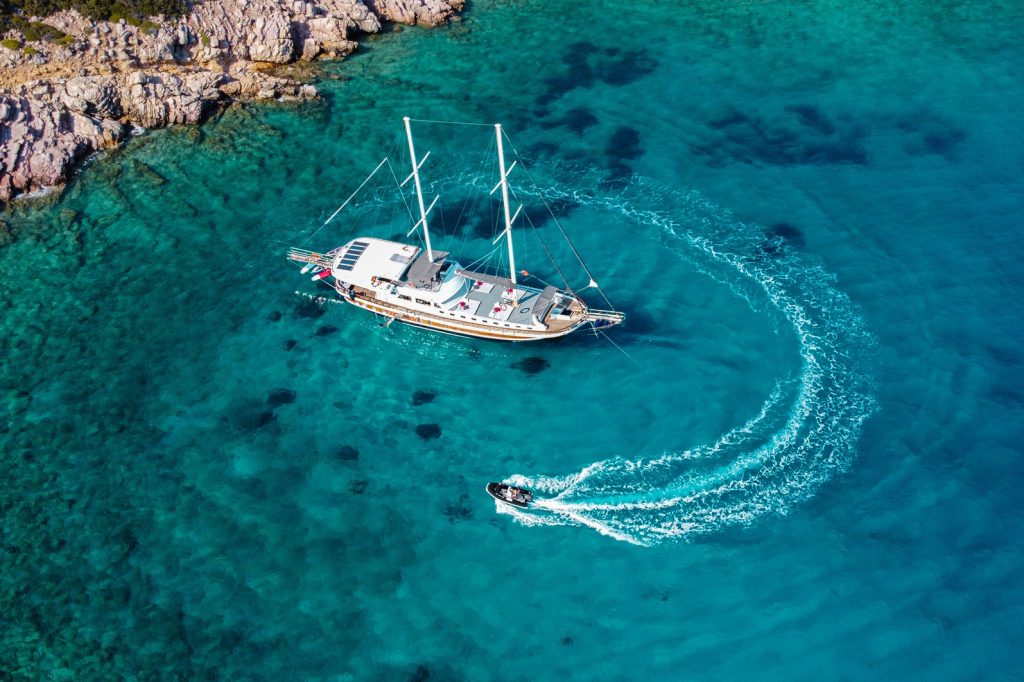 Aerial view of a large sailboat anchored in clear turquoise water near a rocky shoreline, with a smaller motorboat making a circular wake nearby.