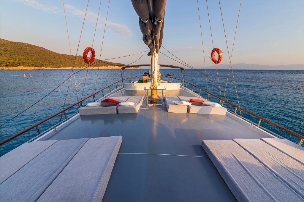 View from the deck of a luxury sailboat with cushioned lounge chairs and life rings, floating on calm blue water near a coastline with green hills under a clear sky at sunset.
