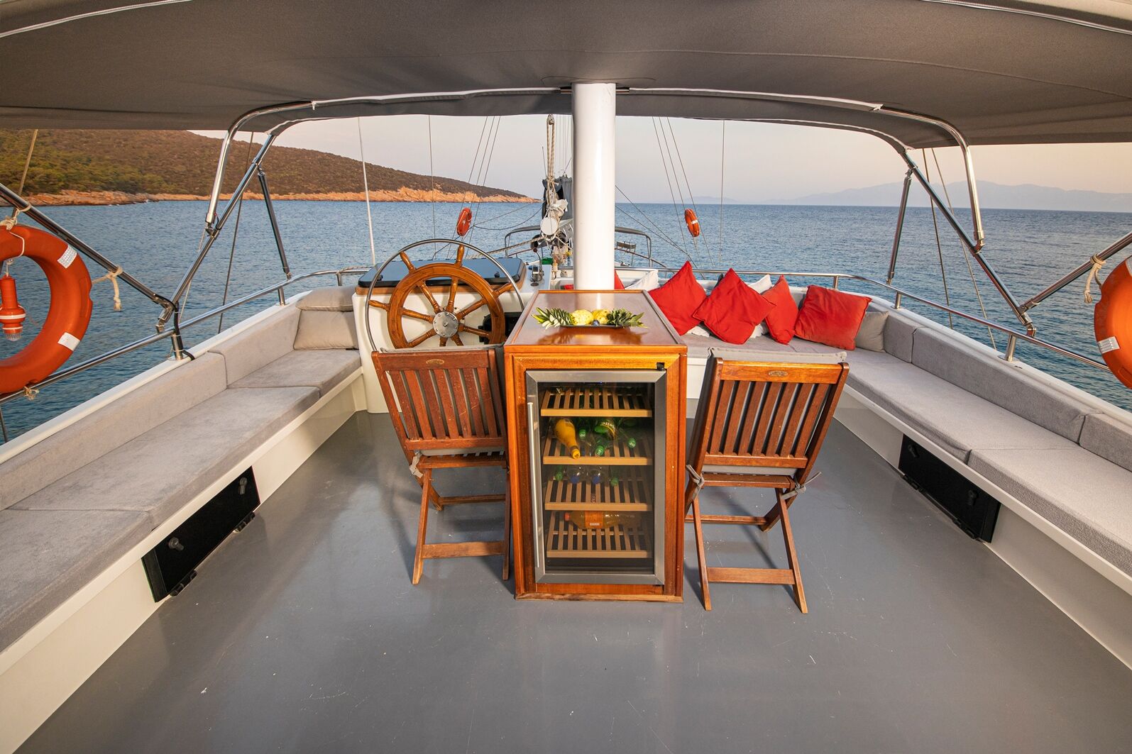 The deck of a yacht with wooden chairs, a small fridge, red cushions, a wooden steering wheel, and views of calm water and distant hills under a canopy.